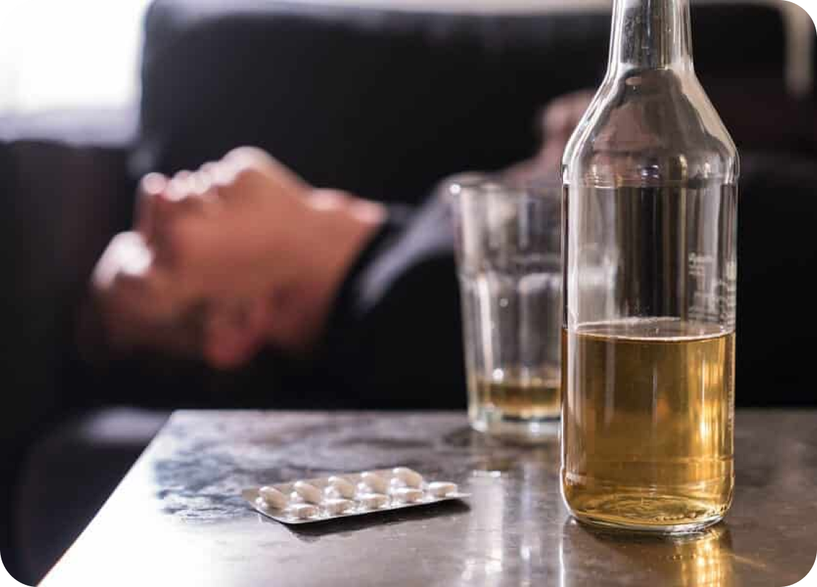 Man in the back laying on a couch with alcohol and pills on the table in front of him